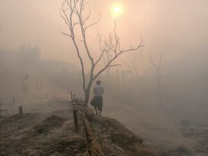 Image description: A pink mist covers the burnt remains of a Rohingya camp. Even the trees stand leafless, as if in mourning. Two people can be seen against the fading light in a landscape that is unsettling, mixing beauty with sadness. March 11, 2023